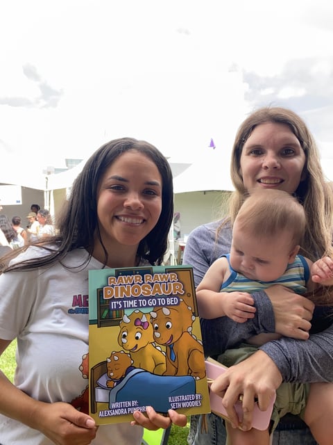 Two women and a toddler smiling outdoors while holding a childrens book titled Rawr Rawr Dinosaur.