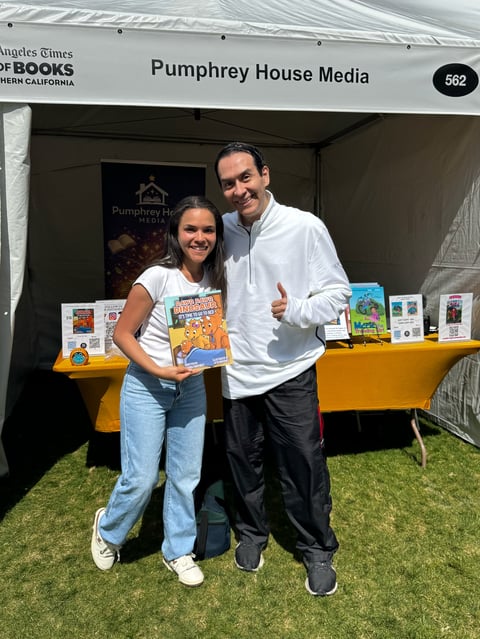 Two people standing at Pumphrey House Media booth at a book festival, with one holding a childrens book and yellow tables displaying published works behind them