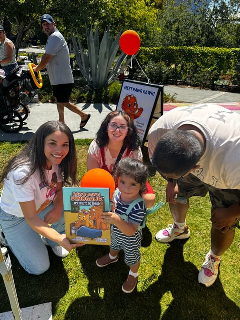 Two women and a young child holding a childrens book at an outdoor community event in a grassy park setting