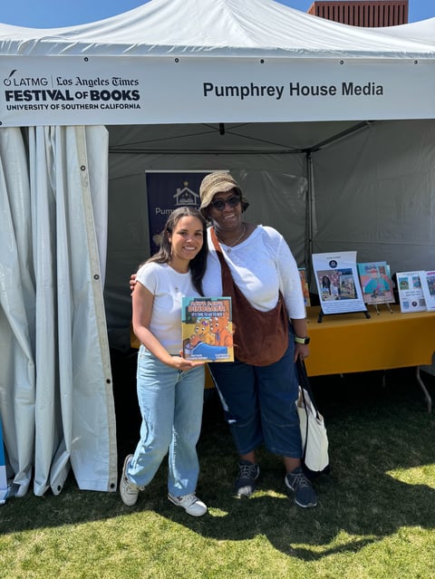 Two people at Pumphrey House Media booth at LA Times Festival of Books, woman holding book, man in hat, white tent with signage visible