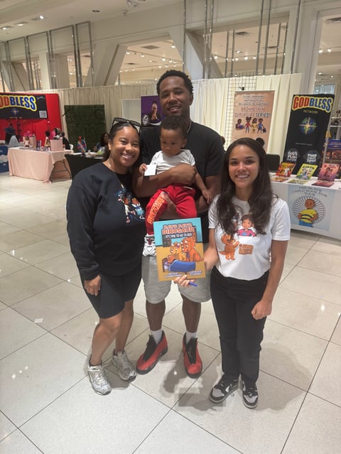Family of four posing at what appears to be a convention or event, holding comic books and merchandise in an indoor venue with booths in the background