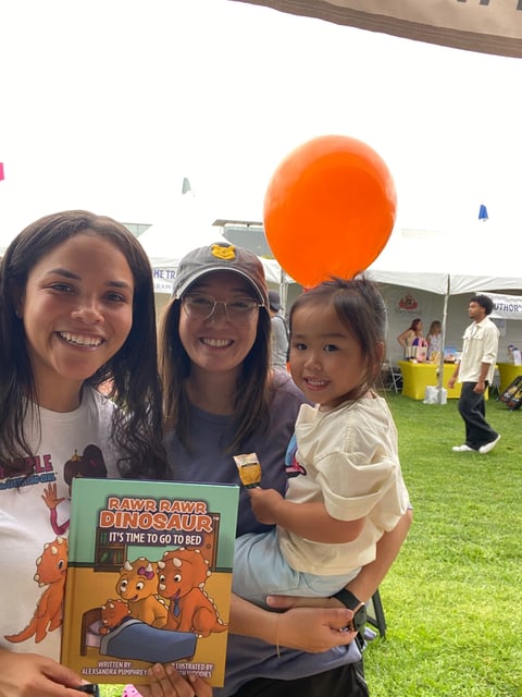 Three smiling girls holding a childrens book and orange balloon at an outdoor event on a grassy field