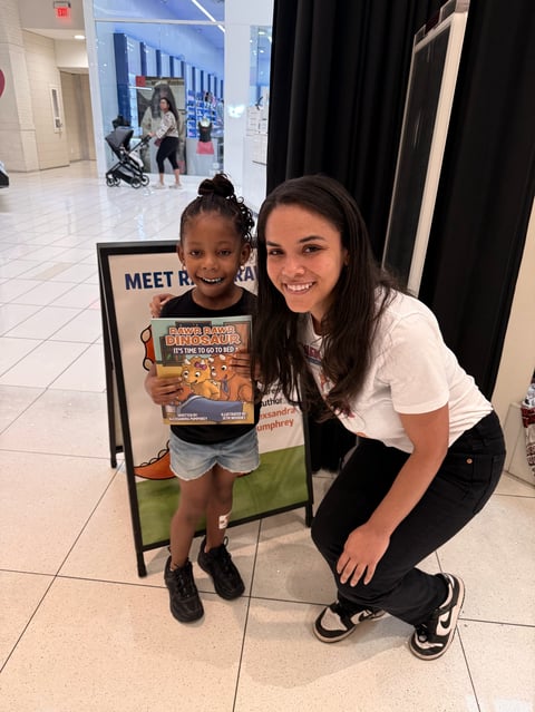 Young girl and teen girl smiling at camera holding childrens book at indoor event with meet and greet signage