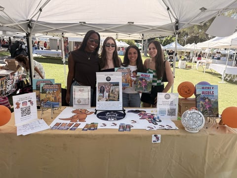 Four women standing behind a vendor booth at an outdoor event displaying artwork, prints, and merchandise under a tent