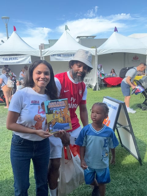 Three people at an outdoor festival holding childrens books under white tents with other visitors and activities in the background