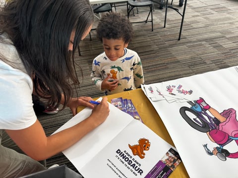 Woman and young child coloring dinosaur and vehicle drawings at a table together