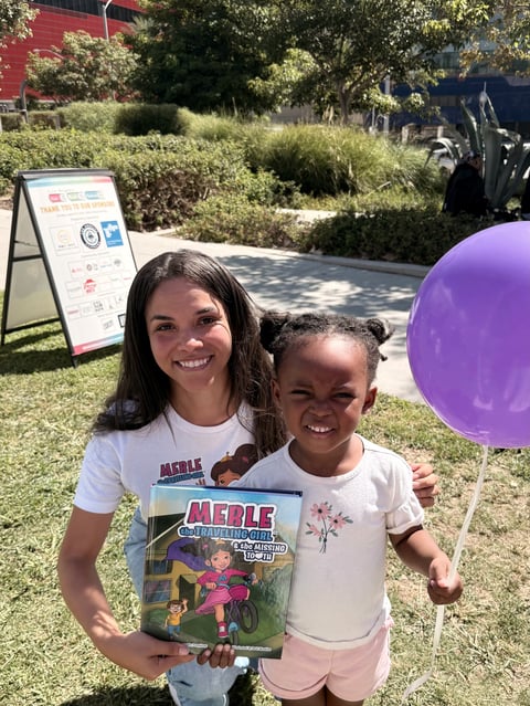 Two young girls smiling at an outdoor event, one holding a purple balloon and a Merle book