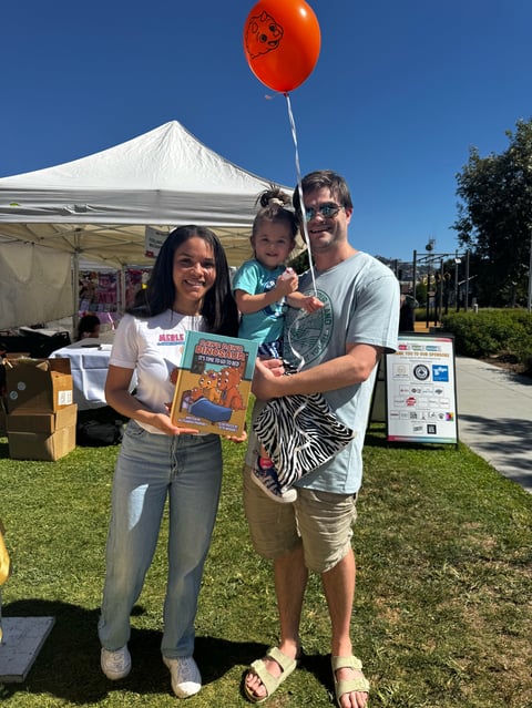 Family of three at outdoor event holding books and red balloon under white tent with clear blue sky