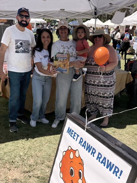 Five people smiling at an outdoor event under a tent with a Meet Rawr Rawr sign featuring an orange character icon visible in foreground