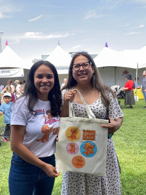 Two young women at an outdoor festival holding a tote bag with cartoon characters, white tents visible in background