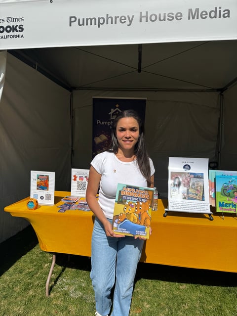 Woman holding a childrens book at Pumphrey House Media booth display with yellow table and promotional materials