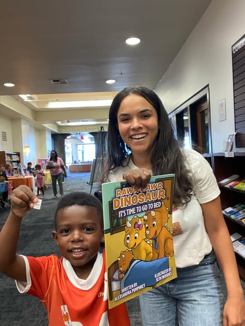 A young woman and small child holding up a Ravi the Dinosaur childrens book in a library setting, both smiling at the camera