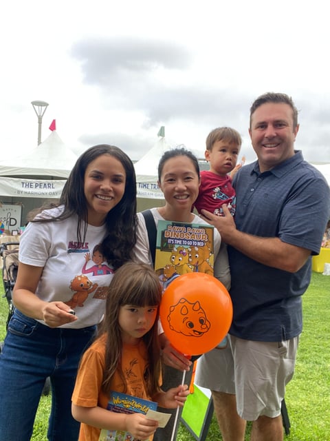 Family of five smiling at outdoor event with children holding orange balloon and dinosaur book