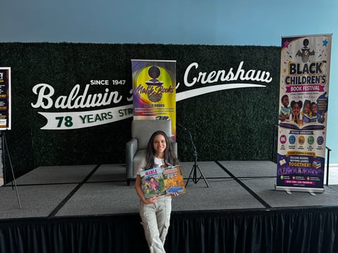 Woman holding magazines standing on stage in front of Baldwin 78 Years and Crenshaw signage with green backdrop