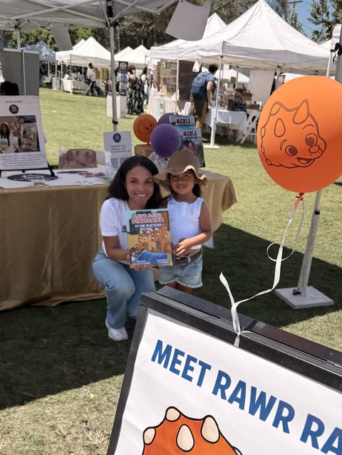 Woman and child holding a book at a Meet Rawr Rawr booth at an outdoor festival with white tents and an orange balloon