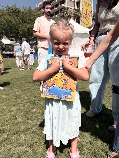 Young girl smiling and holding a comic book at an outdoor event with people in background