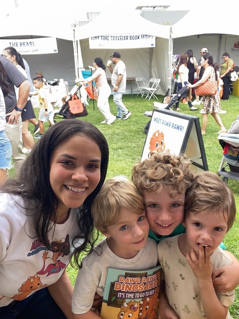 A teenage girl and three young children smiling at an outdoor community event with vendor tents and families in the background
