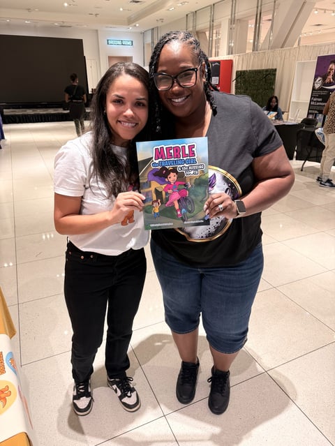 Two women smiling at a convention, holding a Merle action figure toy box, indoors with tiled flooring and modern background