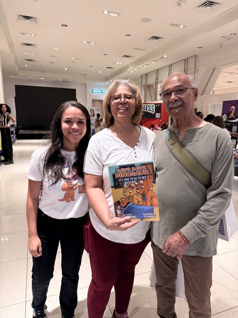 Three people smile at an indoor event while the woman in the center holds a graphic novel or comic book.