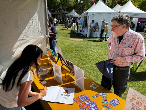 Man in patterned shirt speaking with woman at outdoor yellow booth at community fair with white tents in background
