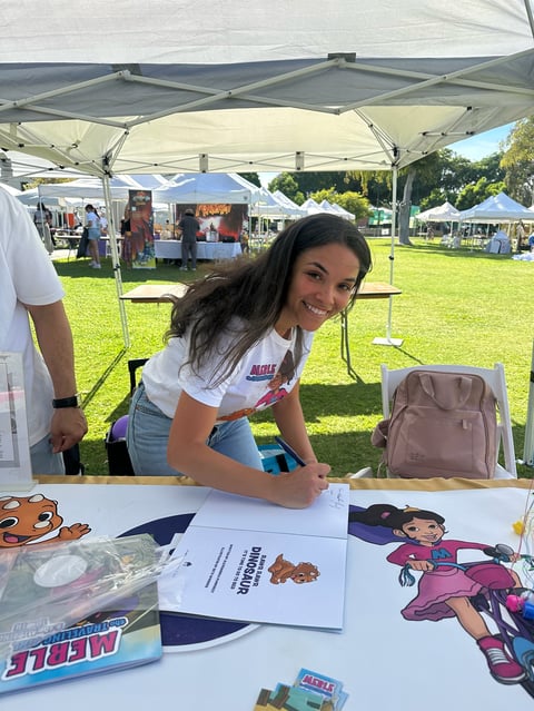 Woman at outdoor market booth under white tent, smiling at camera with merchandise and promotional materials on table