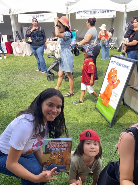 Young woman holding a childrens book at an outdoor event with vendors and families on grass