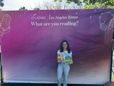 Woman holding a childrens book standing in front of a Los Angeles Times purple billboard asking What are you reading?