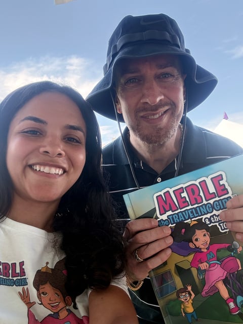 Two people smiling at the camera holding a Marble comic book, outdoors in bright sunlight with blue sky background