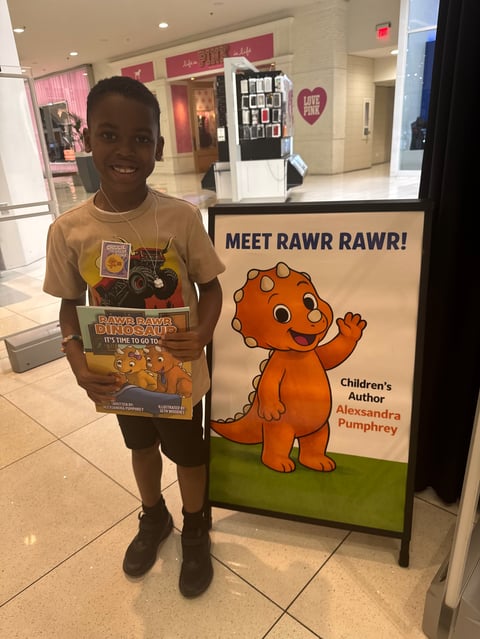 Child smiling beside a promotional sign for the Rawr Rawr character by children's author Alexsandra Pumphrey in a mall