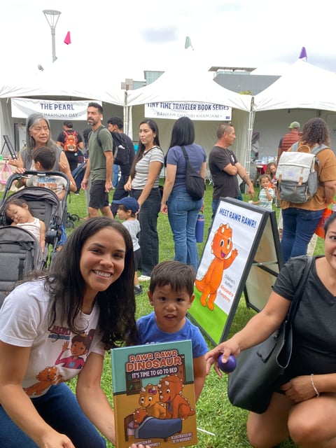 Woman and child at outdoor book fair event holding a dinosaur-themed childrens book among crowds and vendor tents