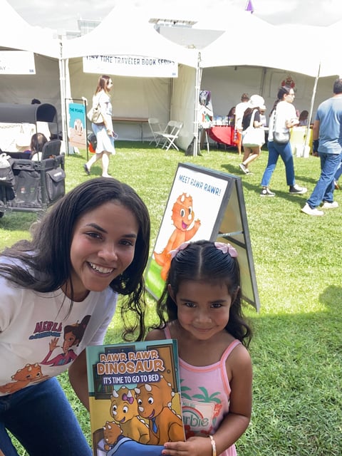 Two smiling girls holding childrens books at an outdoor event with tents and people in the background