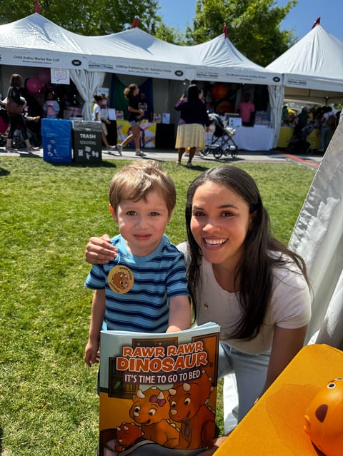 Young boy and woman smiling at outdoor event holding medal and dinosaur-themed sign with white tents in background