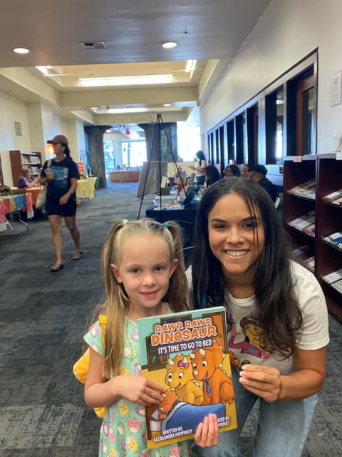 Two girls smiling at camera while holding a Rawr Ava Dinosaur childrens book in a library
