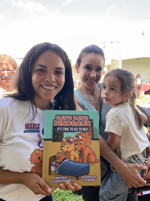 Three women and a young girl holding a childrens book titled Rawr Rawr Dinosaur, smiling at an outdoor event.