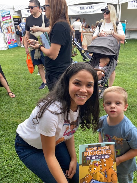 Young woman and toddler boy smiling at outdoor event, holding childrens dinosaur book, with festival tents and families in background